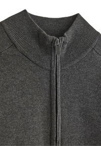 Tmavě šedý pletený svetr s vroubkovaným vysokým límcem a středovým stříbrným zipem, částečně rozepnutý.