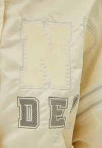 Kremowa satynowa kurtka z dużymi haftowanymi literami "N" i "DE" z teksturami oraz subtelnymi niebieskimi akcentami. Lekka i gładka w dotyku.