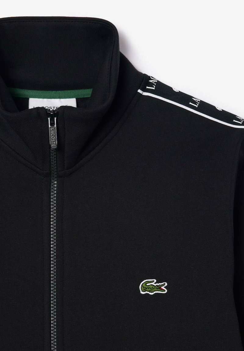 Schwarze Zip-Jacke aus glattem Material, mit stehendem Kragen, grünen Akzenten und einem kleinen gestickten Krokodil-Logo auf der Brust.