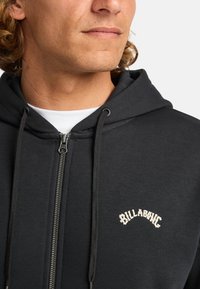 Schwarze Zip-Up-Hoodie mit verstellbaren Kordeln und weißem, gesticktem "BILLABONG"-Logo. Glatte Stofftextur; Nahaufnahme des Kragens.