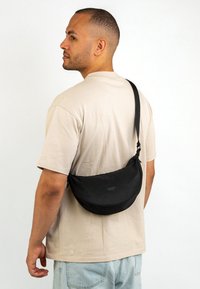 Uomo che indossa una maglietta beige chiaro e jeans blu chiaro con una borsa a tracolla nera appesa dietro la schiena, su uno sfondo bianco semplice.