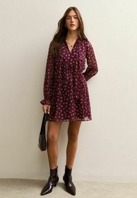 Vestido burdeos con escote en V, mangas largas transparentes, patrón floral, cintura ajustada y falda acampanada, combinado con botas negras hasta el tobillo y un bolso.