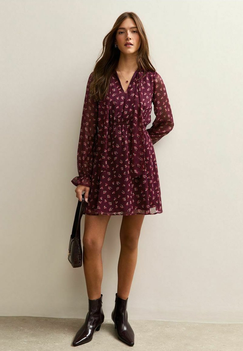 Vestido burdeos con escote en V, mangas largas transparentes, patrón floral, cintura ajustada y falda acampanada, combinado con botas negras hasta el tobillo y un bolso.