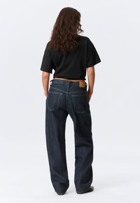 Σκούρα μπλε τζιν από denim με άνετη γραμμή και σχεδίαση με πέντε τσέπες· διαθέτουν ένθετο από καφέ δέρμα στη ζώνη πίσω και κίτρινες ραφές ως διακοσμητικά στοιχεία.