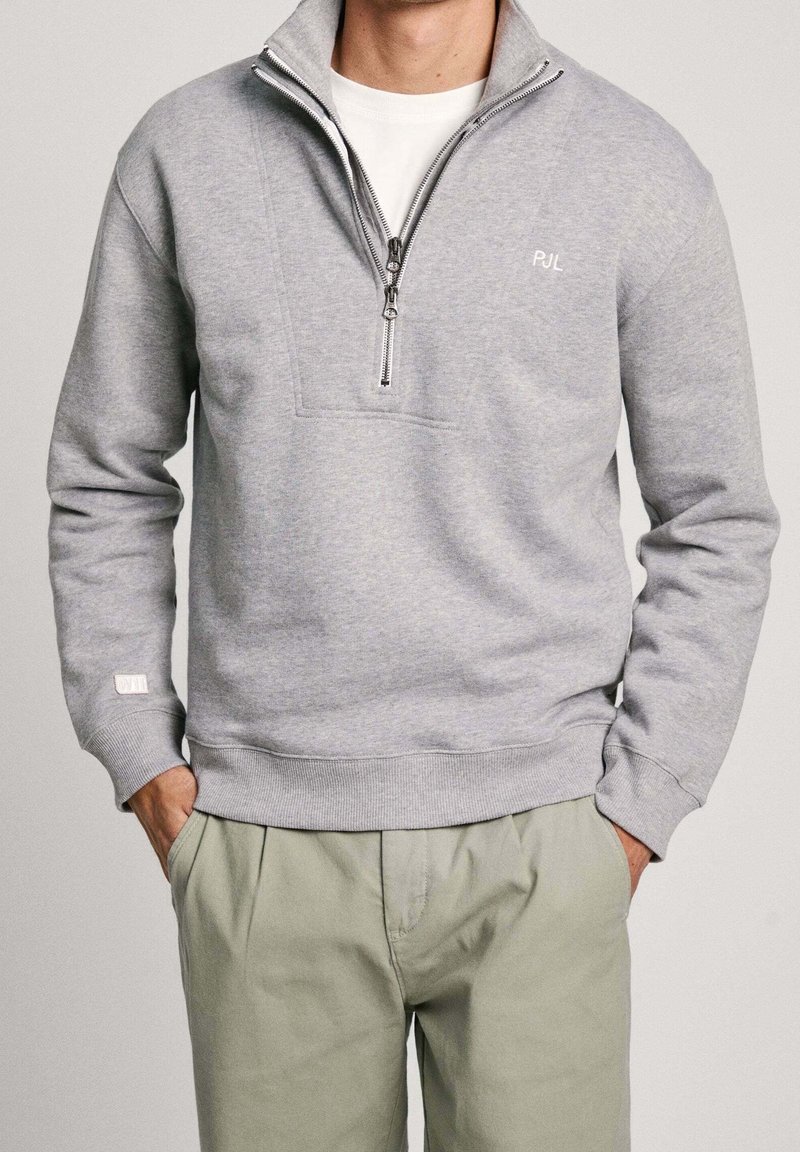 Felpa grigia con zip e collo alto, presenta una cucitura sul petto e un piccolo logo bianco. Maniche con polsini e vestibilità comoda.