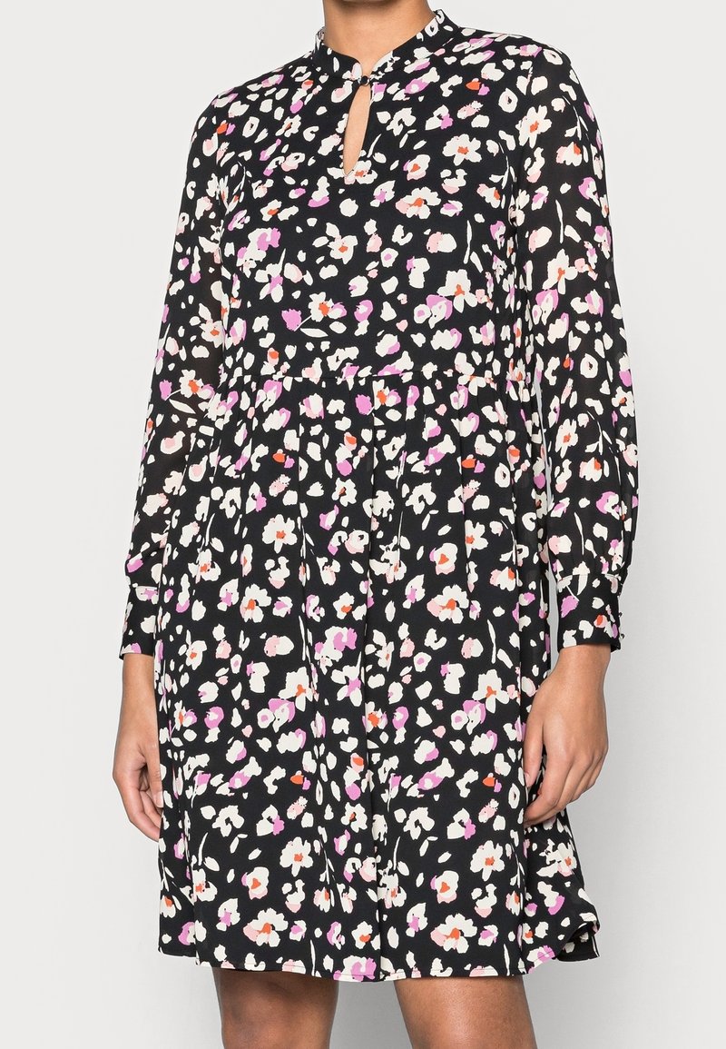 Robe noire à manches longues avec imprimé floral rose, blanc et orange, encolure américaine et taille froncée portée par une personne debout.
