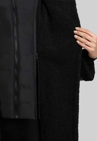 Sort overtøj med et struktureret, fluffy materiale og et glat, polstret indre lag. Lynlåsdetalje synlig langs midten foran.