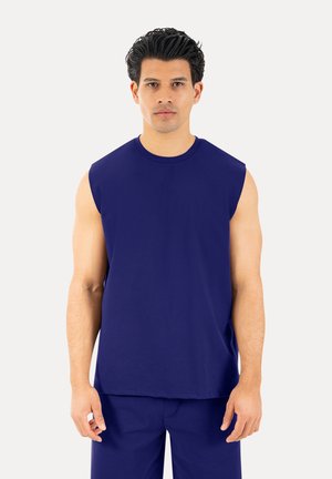 Hombre con cabello corto y oscuro que lleva una camiseta sin mangas azul marino lisa y pantalones a juego, de pie sobre un fondo blanco.