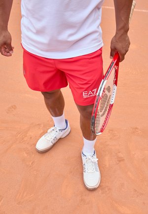 Pantaloni sportivi rossi con logo, t-shirt bianca e scarpe da tennis bianche con accenti blu. Tiene una racchetta da tennis rossa su un campo in terra rossa.