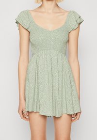 Femme portant une robe fleurie courte de couleur vert clair avec des manches volantées et une taille cintrée, se tenant devant un fond blanc uni.