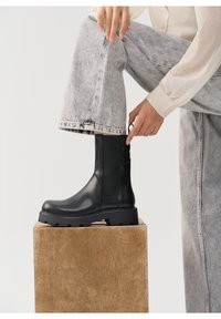 Botas de cuero negras hasta el tobillo con una suela gruesa, paneles laterales elásticos y una lengüeta, combinadas con jeans de pierna ancha en gris claro.