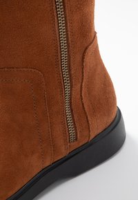 Bruine suède enkellaars met een rits aan de zijkant, zwarte rubberen zool, subtiele textuur en zichtbare stiksels.