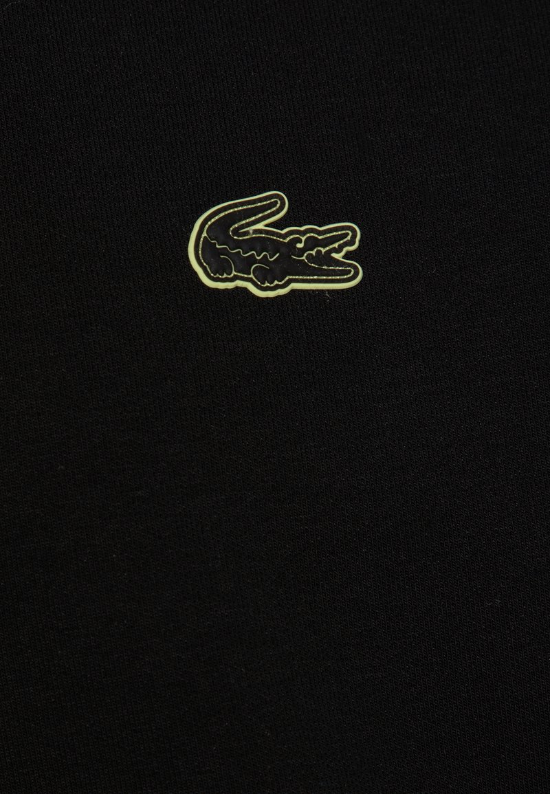 Tissu en coton noir avec un petit logo de crocodile en relief, accentué de vert, brodé sur le côté gauche. Surface texturée, design simple.