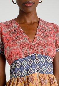 Robe à motifs multiples avec des motifs floraux rouges et bleus. Décolleté en V, manches courtes bouffantes et détails en boutons à la taille. Tissu texturé.