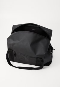 Svart duffelbag laget av slitesterkt materiale, med glidelås på toppen, en frontlomme og justerbare stropper for bæring.