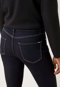 Jean en denim bleu foncé avec des coutures orange, comportant des poches arrière et un design slim. Le tissu a une texture lisse et une forme structurée.
