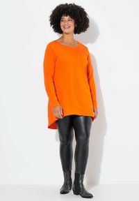 Langarmige oranje tuniek met een ronde hals, gecombineerd met zwarte neplederen leggings en enkellaarzen met zilveren studs.