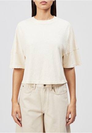 Frau trägt ein lockeres, kurzärmeliges beige Crop-Top und hoch taillierte beige Hose, steht vor einem einfarbigen Hintergrund.