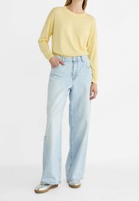 Un pull jaune à manches longues, un jean large bleu clair avec des poches avant, et des chaussures argentées avec un léger talon. Tenue décontractée avec un minimum de déchirures.