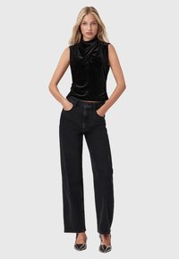 Blonde vrouw in een mouwloze zwarte fluwelen top, hoge, losse zwarte jeans en zwarte puntige hakken, staande met handen in de pockets.