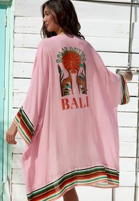 Vaaleanpunainen kimono-tyylinen haalari, jossa on raidallisia vihreitä, oransseja ja valkoisia yksityiskohtia. Sisältää kuvan kirahveista ja auringosta, jossa lukee "SOLAR DEITIES BALI".