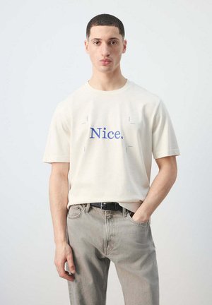 Hombre con el pelo corto, vistiendo una camiseta color marfil con el texto "Nice.", jeans gris claro, de pie frente a un fondo liso y claro.