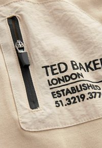 Béžová tkanina so zipsovým vreckom a čiernym kovovým príslušenstvom. Na čiernej potlači je napísané "Ted Baker London Established 51.3219.377."