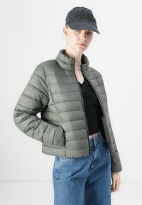 Giacca puffer verde oliva con design trapuntato, colletto alto e chiusura con zip, abbinata a una maglietta nera e jeans a gamba larga blu.