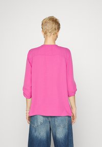 Helles pinkes Blusenshirt mit Dreiviertelärmeln, die an den Bündchen gerafft sind. Verfügt über einen runden Ausschnitt und einen strukturierte Stoff. Getragen mit lockeren, ausgebleichten Jeans.