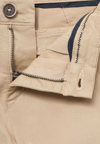 Un tissu beige à la texture lisse présente une fermeture éclair ouverte, mettant en valeur une doublure bleu marine. Un bouton est visible au-dessus de la fermeture éclair.
