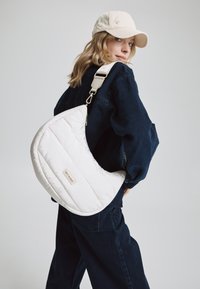 Sac bandoulière blanc matelassé en forme de croissant, avec une sangle beige et une petite étiquette logo. Le modèle porte une tenue en denim bleu foncé.