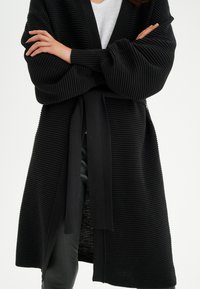 Mustat ribbikuvioiset neulecardiganit, joissa on pitkät hihat ja sidottava vyötärö. Kardigaanissa on teksturoitu pinta ja avonainen etuosa.