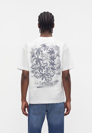 Homme aux cheveux tressés portant un t-shirt blanc avec un imprimé botanique noir et un texte dans le dos, associé à un jean bleu, tournant le dos à la caméra.