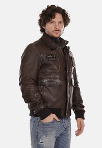 Giubbotto bomber in pelle marrone con zip frontale, polsini a coste e più tasche. Le toppe con simboli sul petto aggiungono un tocco di dettaglio.