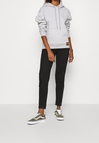 Sudadera gris con capucha y bolsillo frontal, combinada con jeans ajustados negros y zapatillas Vans verde oliva con detalles blancos.