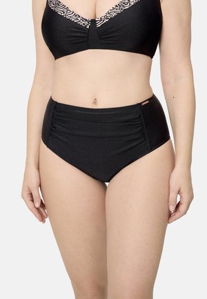 Mujer con un bikini negro con un borde estampado en la parte superior y braguitas de bikini de cintura alta fruncidas, de pie contra un fondo liso.