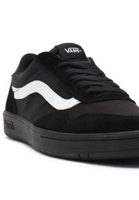 Black sneakers with mesh and suede material, featuring a white wave logo, black laces, and a rubber sole. Minimalist design with padded collar.
