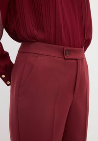 Pantaloni su misura bordeaux con una texture liscia, dotati di tasca laterale e chiusura con bottone in vita.