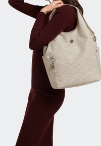 Sac fourre-tout beige à motifs avec une quincaillerie argentée et un charm décoratif, offrant un grand compartiment principal et des poches latérales.