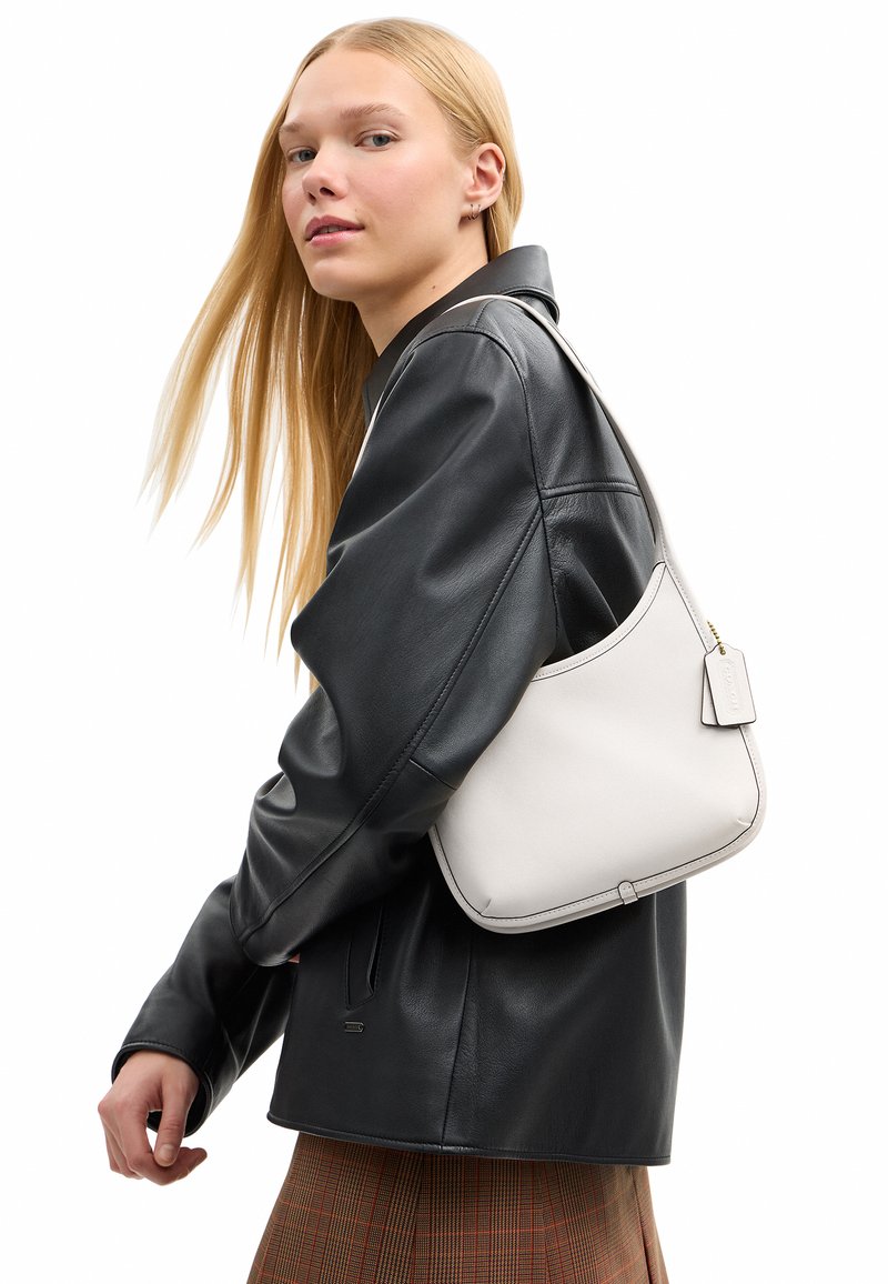Femme blonde portant une veste en cuir noire et une jupe à carreaux, portant un sac à main blanc avec une étiquette.