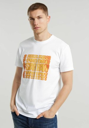 T-shirt bianca in cotone con un bold design di testo in gradiente arancione, con scollo rotondo e maniche corte, indossata con le mani in tasca.