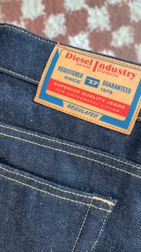 Jeans de ganga azul escura com um rótulo de pele que apresenta texto vermelho e azul. O rótulo indica "Diesel Industry" e "Jeans de Qualidade Superior."