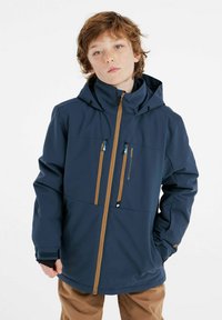 Giacca da esterno blu navy con cappuccio, due tasche a zip sul petto, dettagli delle zip marroni e polsini regolabili. Texture liscia e design sportivo.