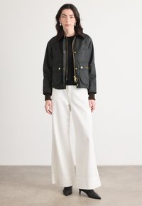 Jachetă neagră ceruită cu un guler din catifea maro, accesorii aurii, două buzunare frontale, purtată peste un top negru și asortată cu pantaloni albi cu croi larg.