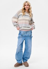 Sweter w paski, dziany w delikatnych pastelowych kolorach, zestawiony z obszernymi spodniami dżinsowymi o unikalnym, skróconym kroju i regulowanych szelkach. Brązowe obuwie.