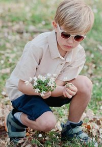 Barn med kort, ljusfärgat hår i vita solglasögon, iförd en beige skjorta och mörka shorts, som håller vita blommor i ett grönområde.