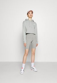 Sweat à capuche gris raccourci avec cordons de serrage et logo, associé à un short de vélo gris. Le mannequin porte des baskets blanches et une casquette rose clair.