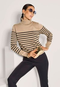 Frau trägt einen beigen gestreiften Rollkragenpullover, schwarze Hose, Sonnenbrille und ein goldenes Armband, während sie eine kleine goldene geometrische Clutch hält.