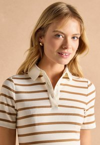 Krémové polo tričko s horizontálnymi hnedými pruhmi, rebrovanou textúrou, krátkymi rukávmi a trojitém gombíkovom zapínaní. Obsahuje klasický golier.