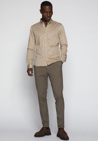 Helle beige Button-Up-Bluse mit langen Ärmeln, kombiniert mit schmalen beigen Hosen und braunen Schnürschuhen. Schlichtes Design, glatte Textur.